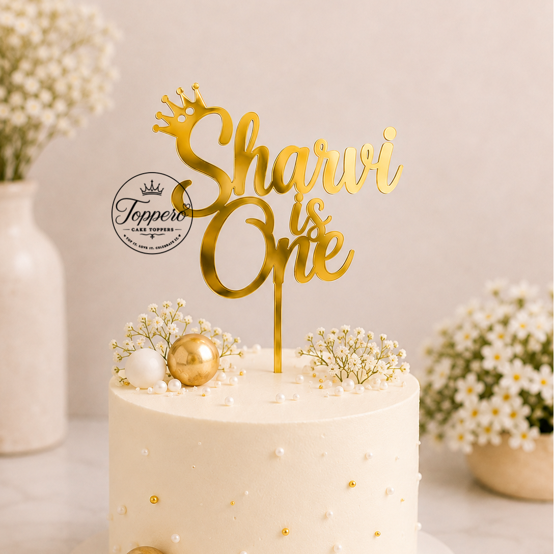 A frosted cream cake on a white board with a gold 'Sharvi is One' acrylic cake topper, decorated with gold and white ornaments and baby's breath flowers. The background features a soft beige wall with blurred floral arrangements.