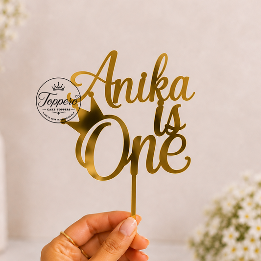 A hand holds a gold metallic cake topper with the script text 'Anika is One' and a small crown icon. The background is a soft beige wall with blurred white flowers in vases.