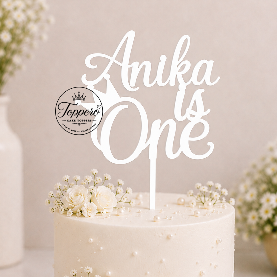 A white frosted birthday cake with pearl-like decorations, a large cream satin bow, and floral accents, topped with a white acrylic cake topper that reads 'Anika is One'. The cake sits on a pink-rimmed white cake board in a softly styled indoor setting with blurred flower arrangements in the background.