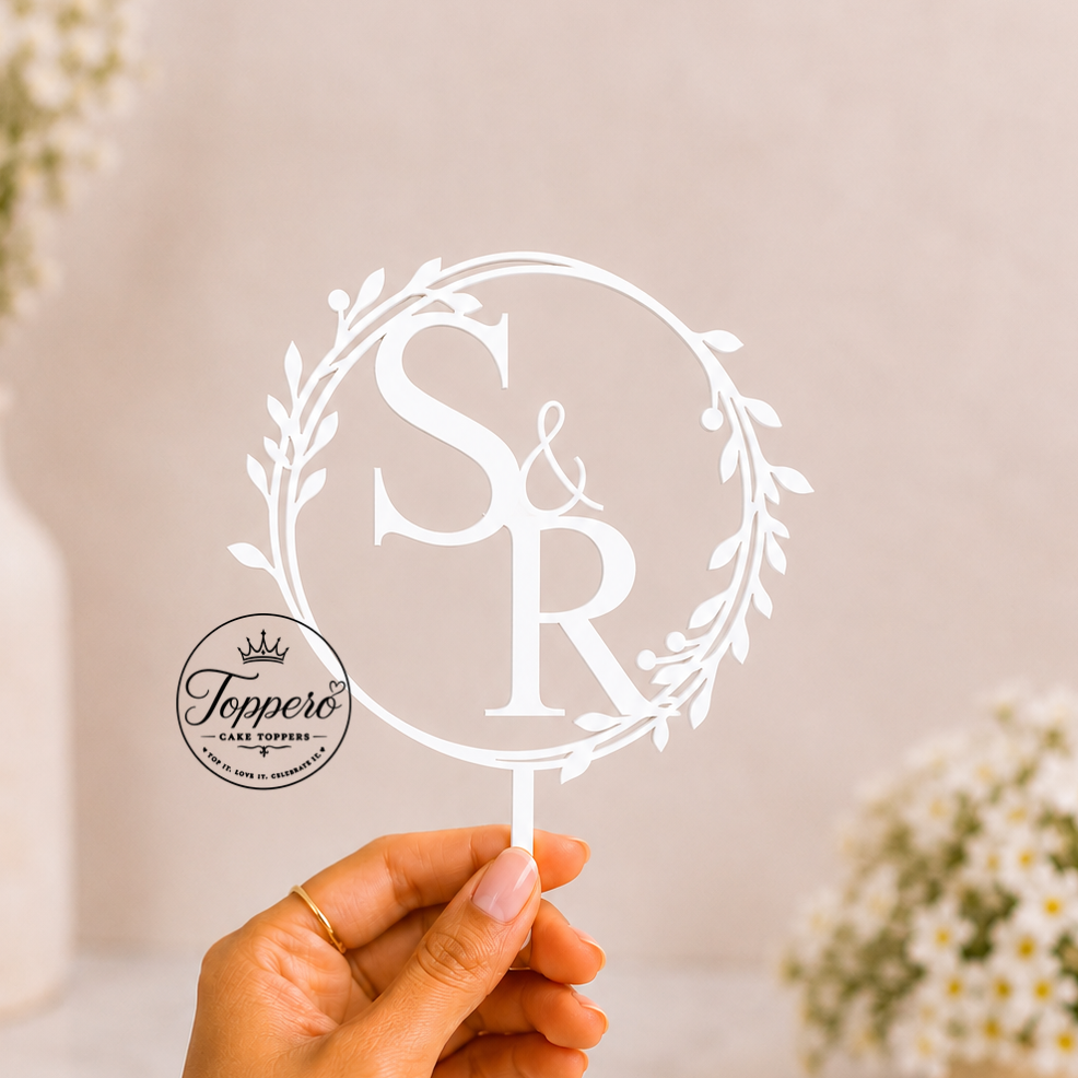 A hand holds a white monogram cake topper featuring the letters 'S & R' inside a leafy wreath. The circular logo for 'Tepper's Cake Toppers' is printed on the product.