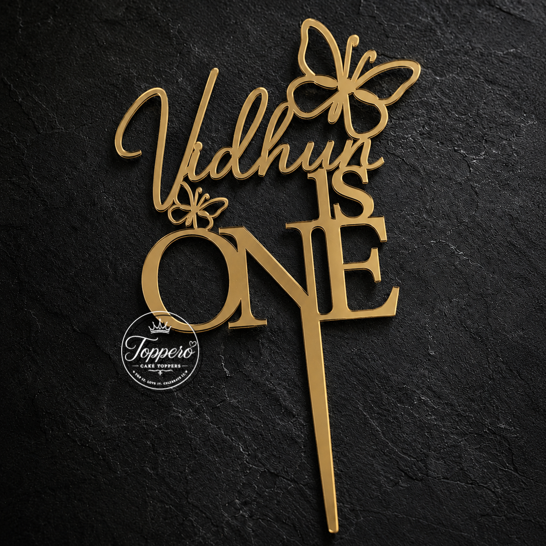 Gold wooden cake topper with the text 'Vidhun IS ONE' and butterfly cutouts, displayed on a dark textured slate background.