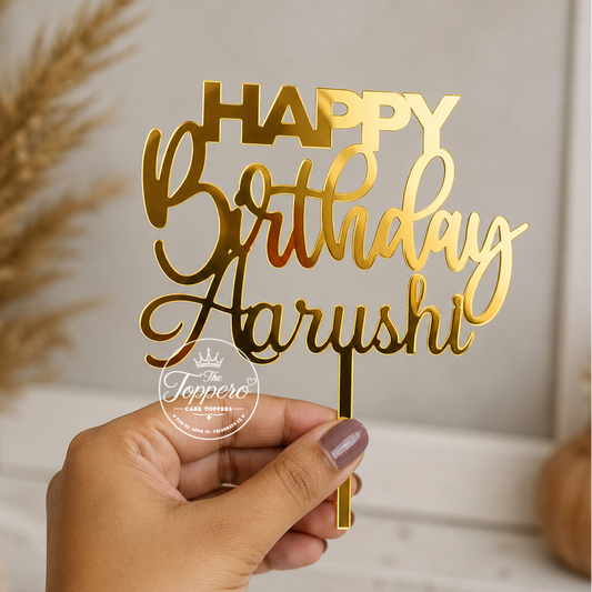 A hand holds a gold metallic laser-cut acrylic birthday cake topper that reads 'HAPPY Birthday Aarushi' against a softly blurred indoor background with dried grass and a white shelf.