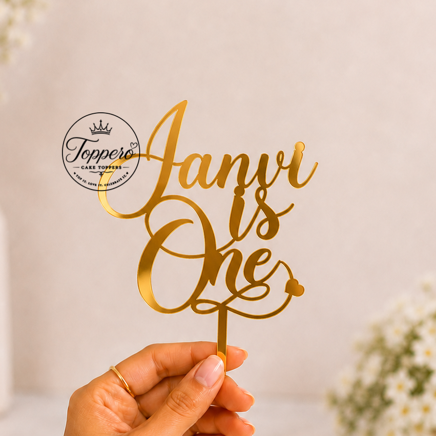 A hand holds a gold metallic laser-cut cake topper that reads 'Janvi is One' against a soft, warm beige background with blurred white flowers.