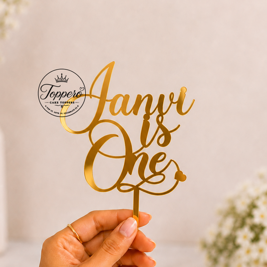 A hand holds a gold metallic laser-cut cake topper that reads 'Janvi is One' against a soft, warm beige background with blurred white flowers.