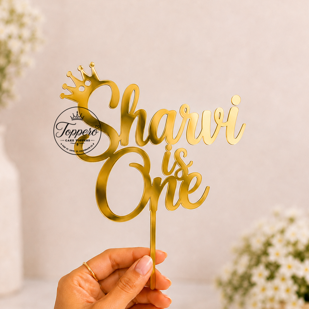 A hand holds a gold laser-cut acrylic cake topper that reads 'Sharvi is One' with a small crown at the top. The background is a soft beige wall with blurred white flowers in vases.
