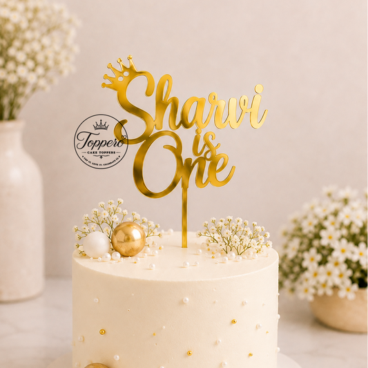 A frosted cream cake on a white board with a gold 'Sharvi is One' acrylic cake topper, decorated with gold and white ornaments and baby's breath flowers. The background features a soft beige wall with blurred floral arrangements.