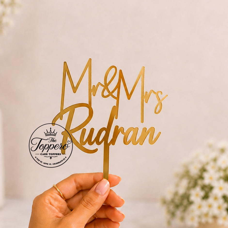 A hand holds a gold metallic script cake topper that reads 'Mr & Mrs Rudran' against a soft, light beige background with blurred white flowers in the background.