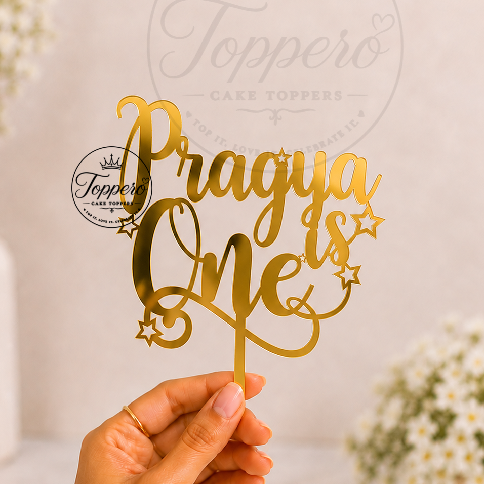 A hand holds a gold metallic laser-cut cake topper that reads 'Pragya is One' with decorative stars, set against a soft-focus background with white flowers and a faint Toppero brand watermark.
