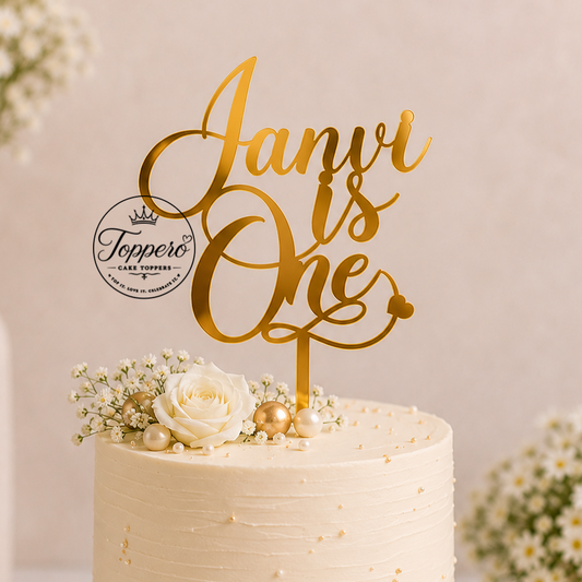 A frosted cream cake on a white and gold round base, decorated with white roses, baby's breath, and pearl-like beads, topped with a gold script cake topper reading 'Janvi is One' and a circular 'Tuppero' logo. The scene is styled with blurred white flower bouquets in the background.