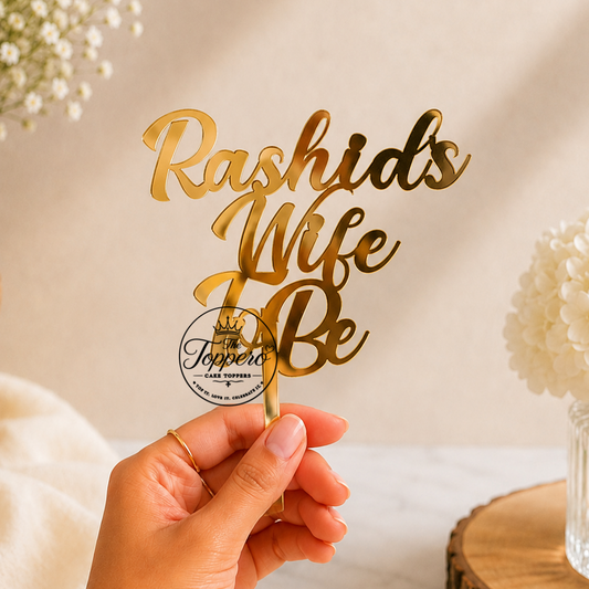 A hand holds a gold metallic script cake topper that reads 'Rashid's Wife Be' against a warm beige background with soft floral props.