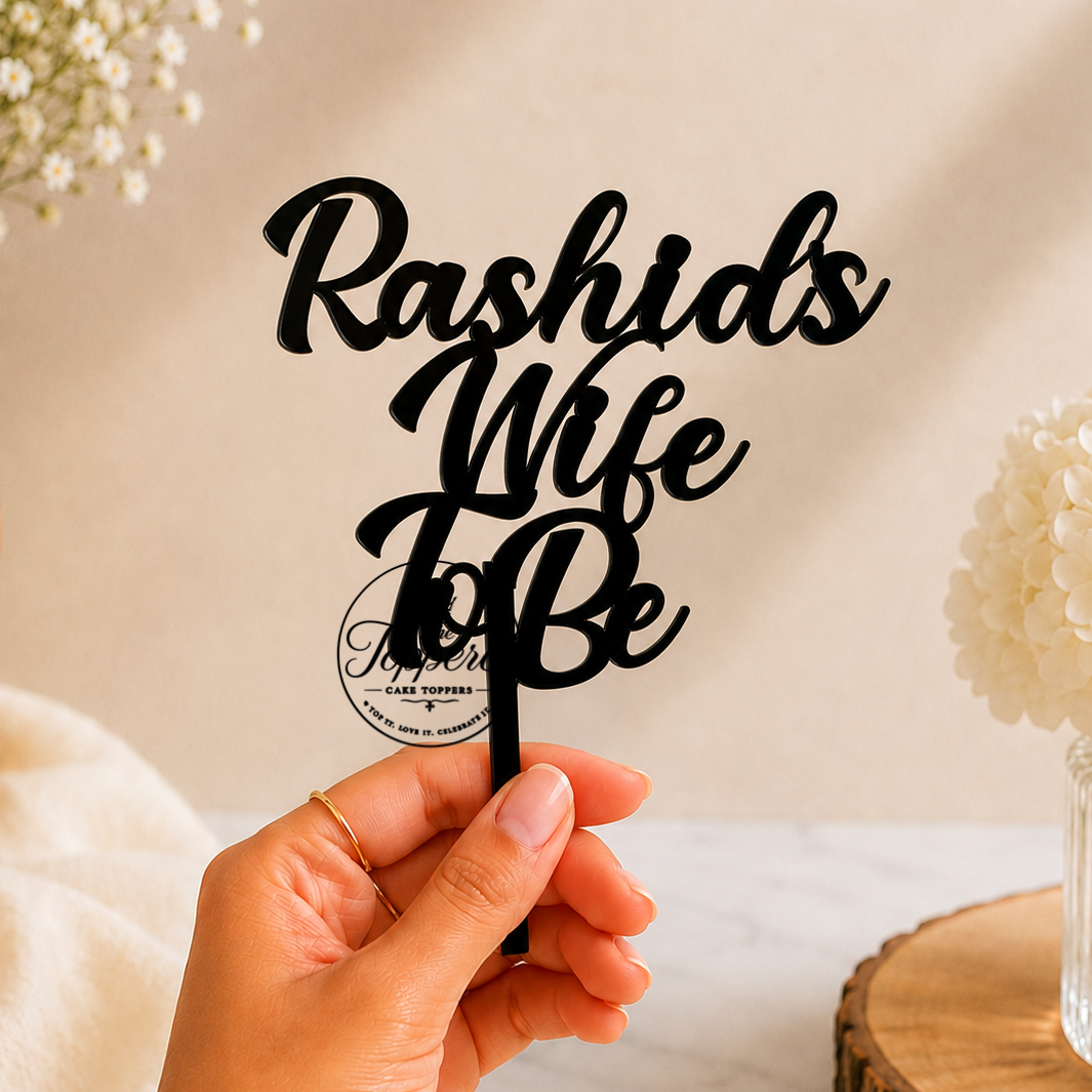 A hand holds a black metal cake topper with the phrase 'Rashids Wife To Be' in elegant cursive lettering, set against a warm beige background with decorative flowers.