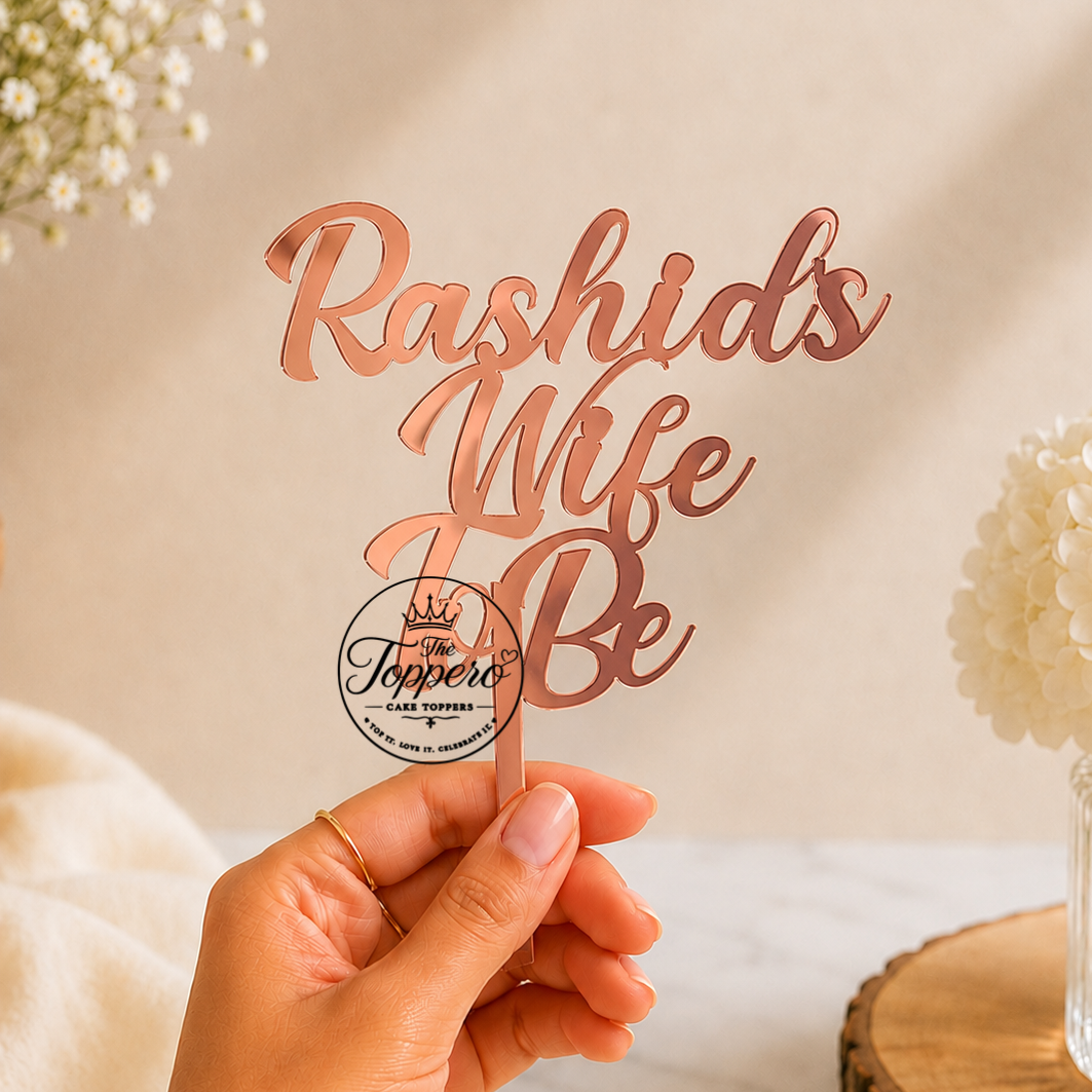 A hand holds a rose gold metallic laser-cut sign that reads 'Rashid's Wife Be' on a wooden stick, with a circular logo for 'The Topper' in the center. The background is a warm beige wall with soft shadows, white flowers in a vase, and a marble surface.