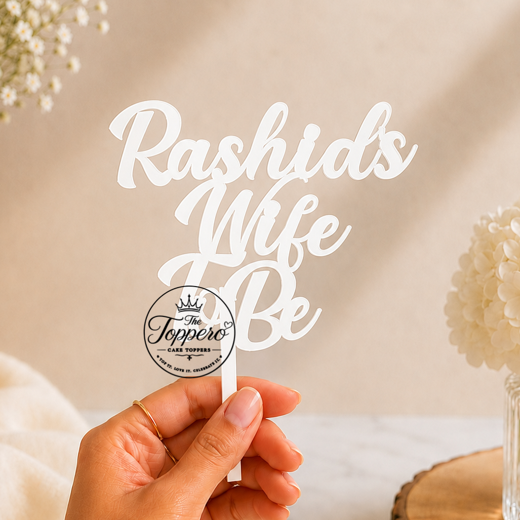 A hand holds a white cake topper with the phrase 'Rashids Wife Be' in elegant script, with a circular logo for 'The Joppeco' visible on the lower part of the topper. The background is a warm beige with soft floral arrangements and a marble surface.