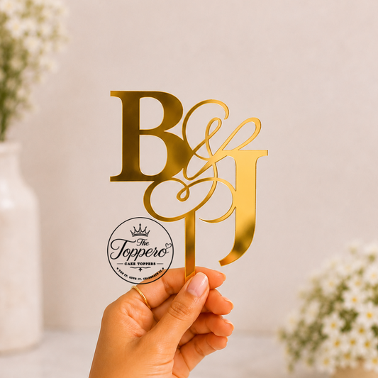 A hand holds a gold monogram table topper with the letters B and J connected by an ampersand, alongside a round logo sticker for 'The Tuppero'. The background is a soft beige with blurred white flowers in vases.