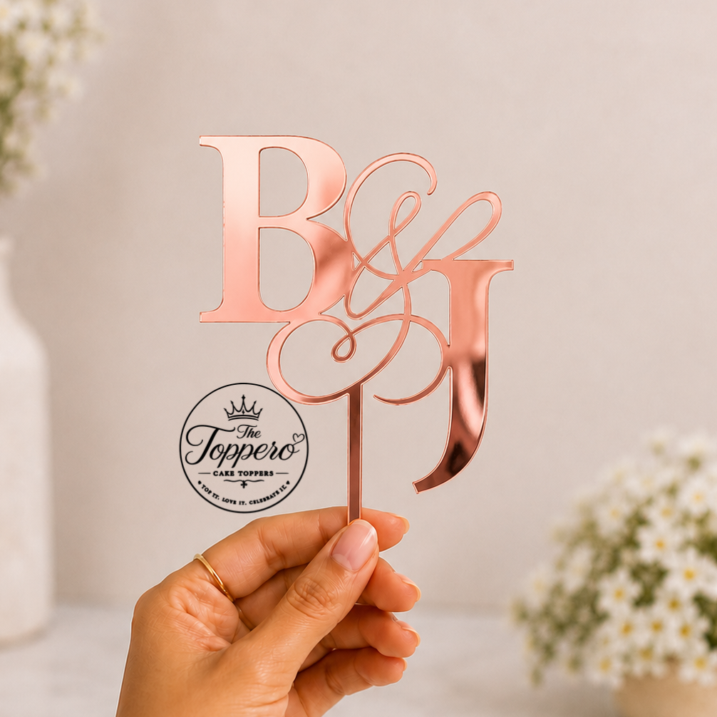 A hand holds a rose gold monogram cake topper featuring the letters B and J with an ampersand, set against a soft-focus background with white flowers.