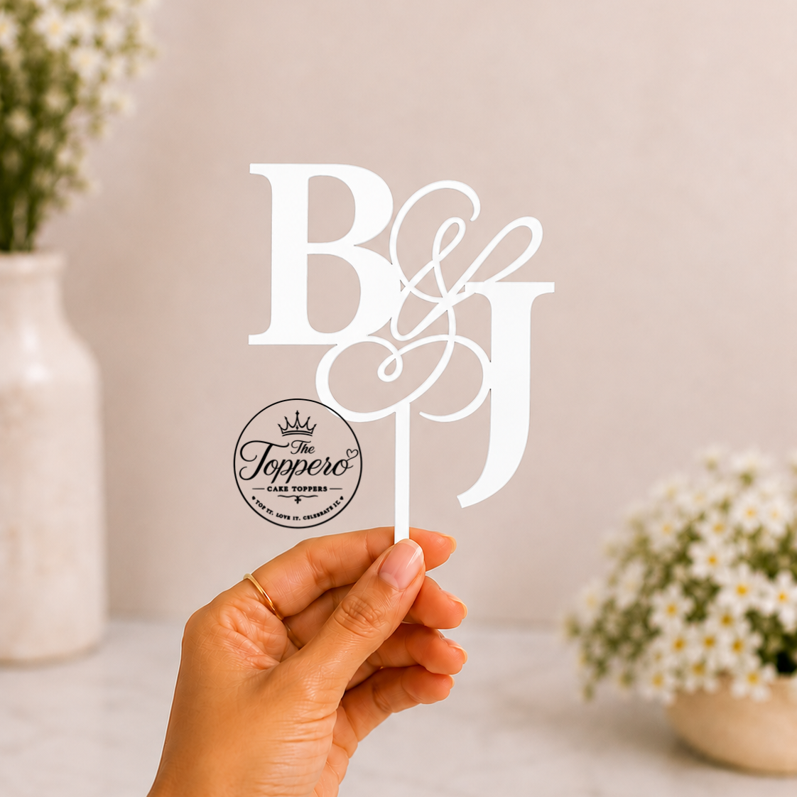 A hand holds a white monogram cake topper featuring the letters B and J with an ampersand, alongside a round black-and-white logo for The Toppo's. The background is a soft beige with blurred white flowers in vases.