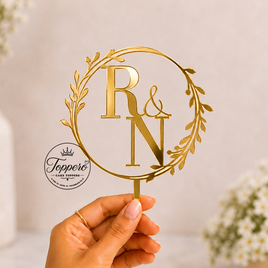 A hand holds a gold metal personalized table topper featuring the letters 'R & N' inside a leafy wreath, set against a soft-focus background with white flowers.