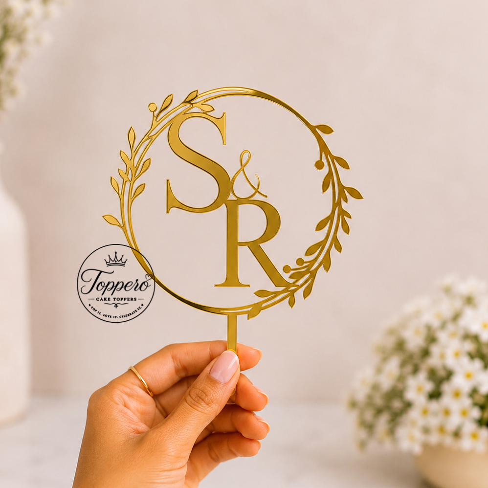 A hand holds a gold metal monogram cake topper featuring the intertwined letters 'S' and 'R' inside a circular wreath design. The background is a soft beige with blurred white flowers in vases.
