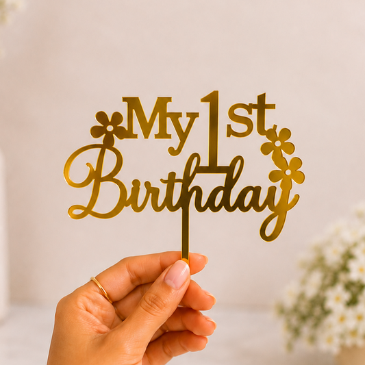 A hand holds a gold metal 'My 1st Birthday' cake topper with small flower accents against a soft beige background with blurred white flowers.
