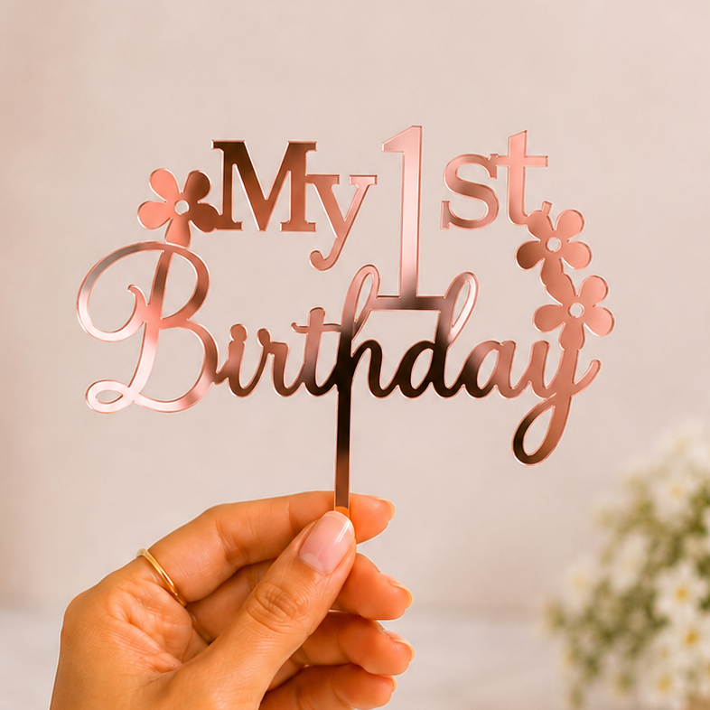 A hand holds a rose gold metal 'My 1st Birthday' cake topper with small flower accents, set against a soft-focus background of white flowers in vases.