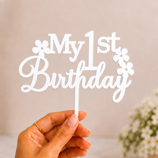A hand holding a white acrylic cake topper that reads 'My 1st Birthday' with small flower details, set against a soft beige background with blurred white flowers in vases.