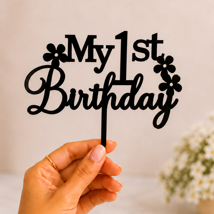 A hand holds a black metal 'My 1st Birthday' cake topper with floral accents against a soft, warm beige background with blurred white flowers.