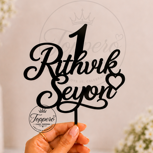 A hand holds a black laser-cut wooden party table topper featuring the text 'Rithvik & Seyon' in an elegant cursive font, with a heart at the end of the word 'Syon' and a large number '1' above it. The background is a soft beige wall with blurred white flowers in vases.