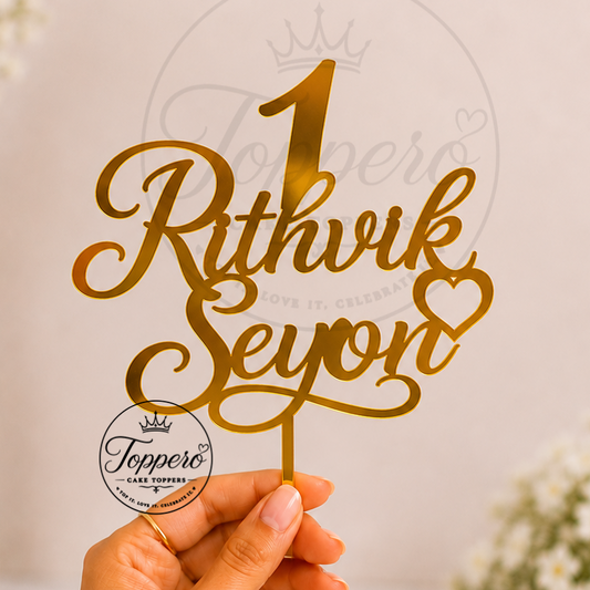 A hand holds a gold metallic laser-cut cake topper with the personalized name 'Rithvik Seyon' and a heart, set against a soft-focus background of white flowers in vases.