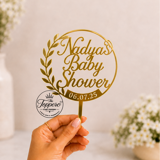 A hand holds a gold personalized baby shower sign with a circular wreath design, reading 'Nadya's Baby Shower' and the date '06.07.