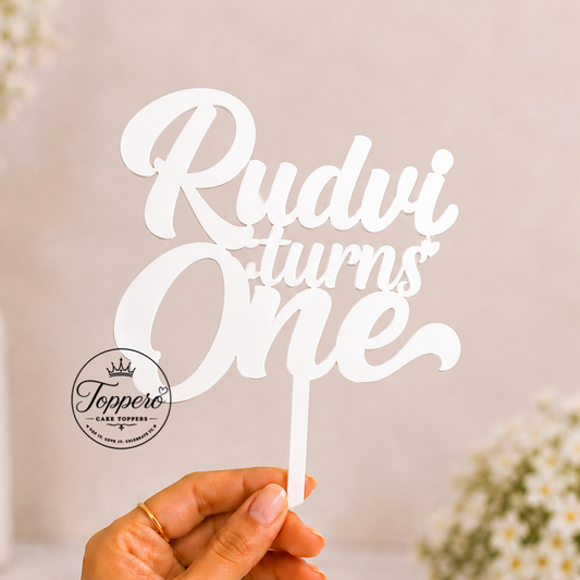 A hand holds a white laser-cut cake topper that reads 'Rudvi turns One' against a softly blurred background with white flowers in vases. A circular logo for 'Teppero' is visible on the topper.
