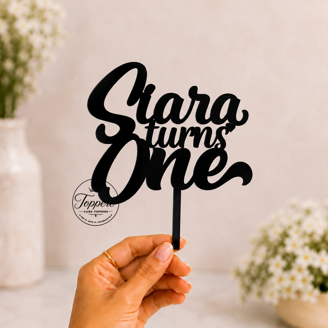 A hand holds a black laser-cut cake topper that reads 'Siara turns One' in elegant cursive and block lettering. The background is a soft, warm beige with blurred white flowers in ceramic vases.