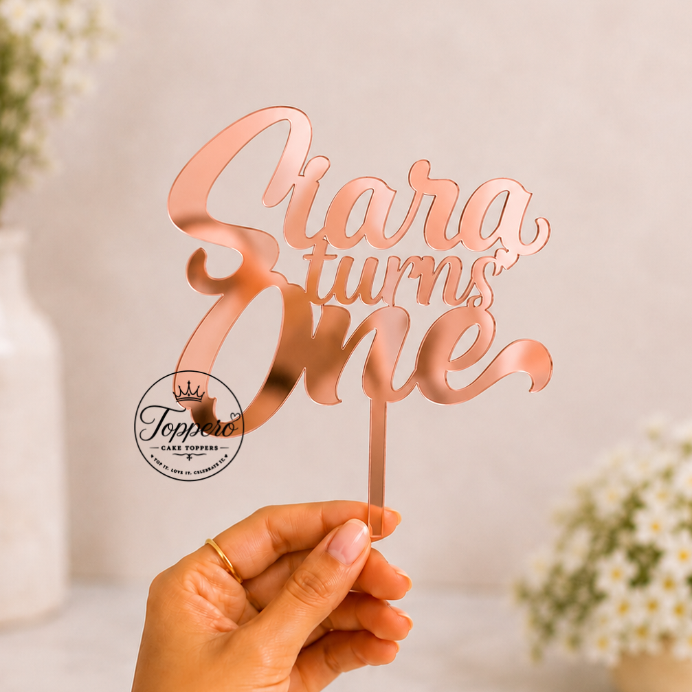 A hand holds a rose gold laser-cut cake topper that reads 'Siara turns One' against a soft-focus background of white daisies in vases.