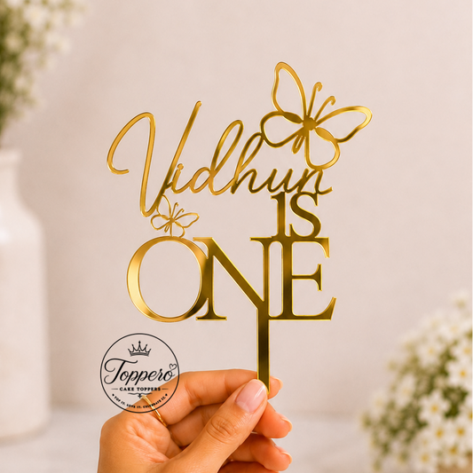 A hand holds a gold laser-cut cake topper with the personalized name 'Vidhvin' and the phrase 'IS ONE' decorated with butterfly and ring motifs, set against a soft, blurred background with white flowers.
