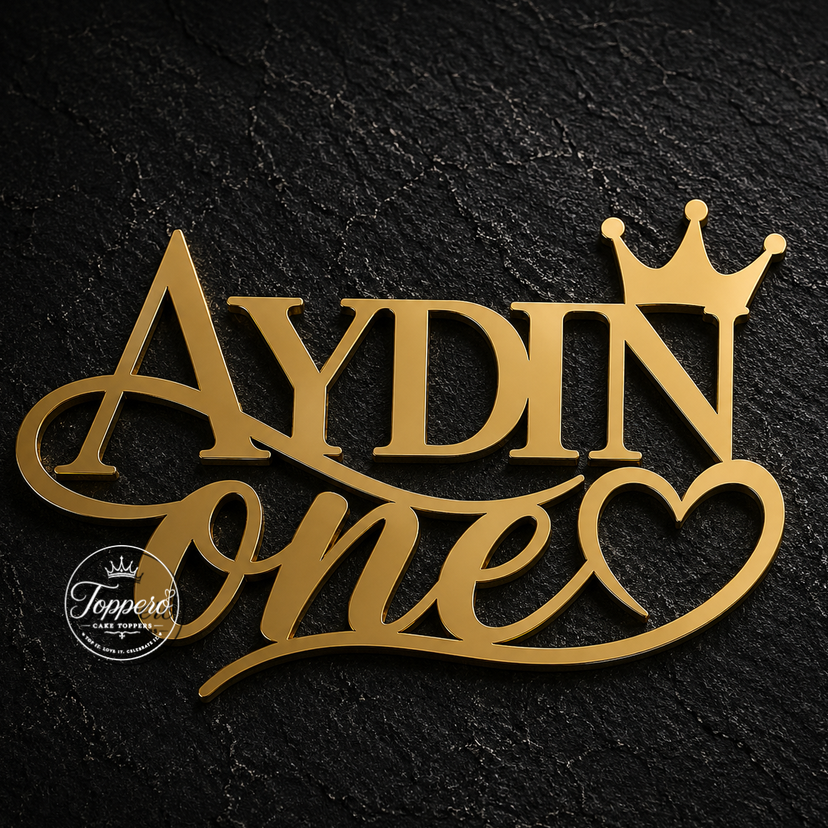 Gold personalized cake topper reading 'AYDIN one' with a small crown and heart, shown on a dark textured background. A circular 'Topers' watermark appears on the lower left.