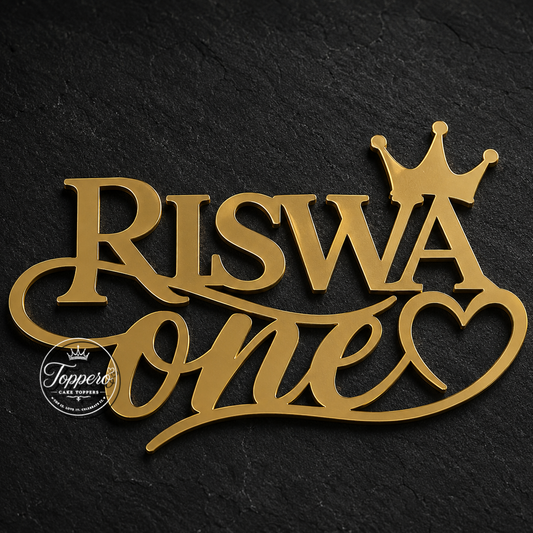 Gold metal cake topper reading 'RISWA one' with a crown and heart, displayed on a dark textured background.