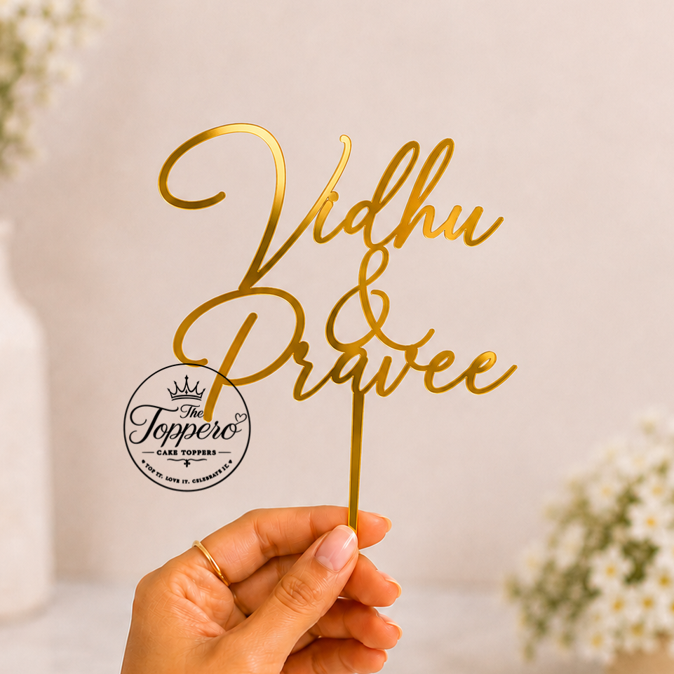 A hand holds a gold metallic script cake topper reading 'Vidhu & Pravee' against a soft, light beige background with blurred white flowers in the background.